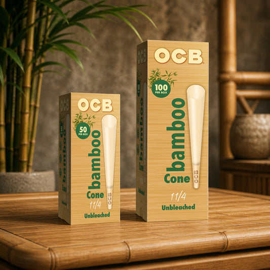 Two boxes of OCB bamboo cone unbleached tobacco, one 50 pack and one 100 pack, displayed on a bamboo table with a blurred background of a potted plant and wooden furniture.