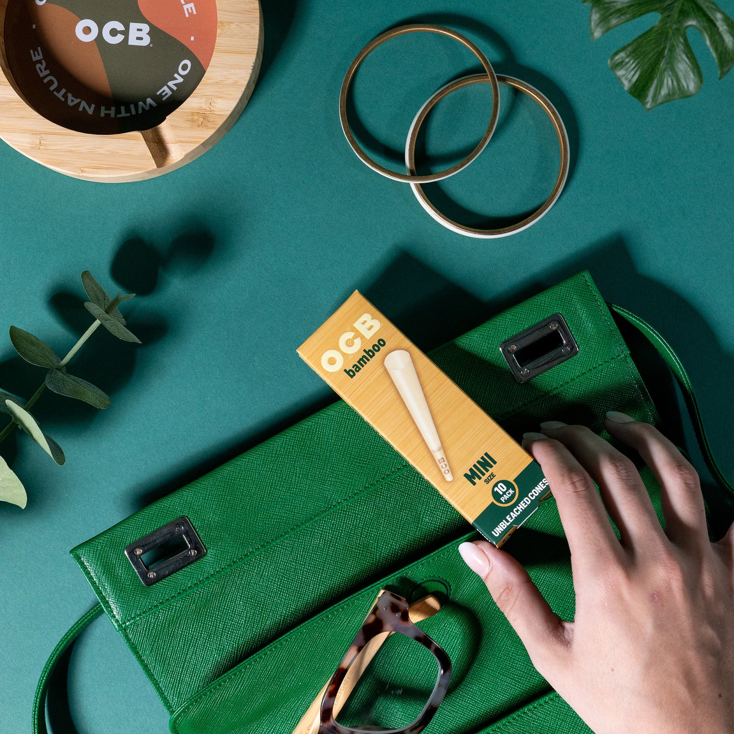 A hand inserts an OCB Bamboo Mini cone pack into a green textured clutch bag, styled on a teal background with gold rings, glasses, and a wooden coaster.