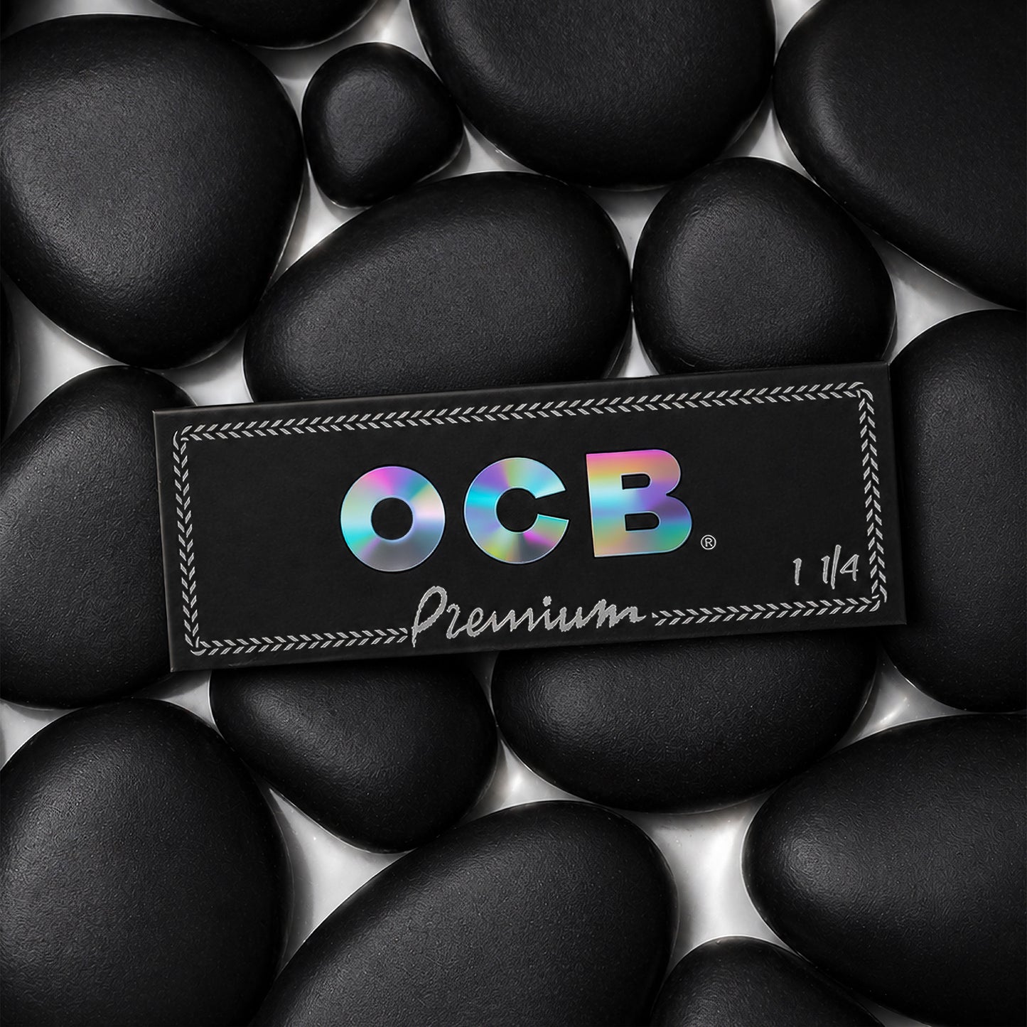 A rectangular black OCB Premium 1 1/4 booklet placed on top of smooth black stones. The OCB logo appears in iridescent rainbow lettering, with 'Premium' and '1 1/4' printed below in white.