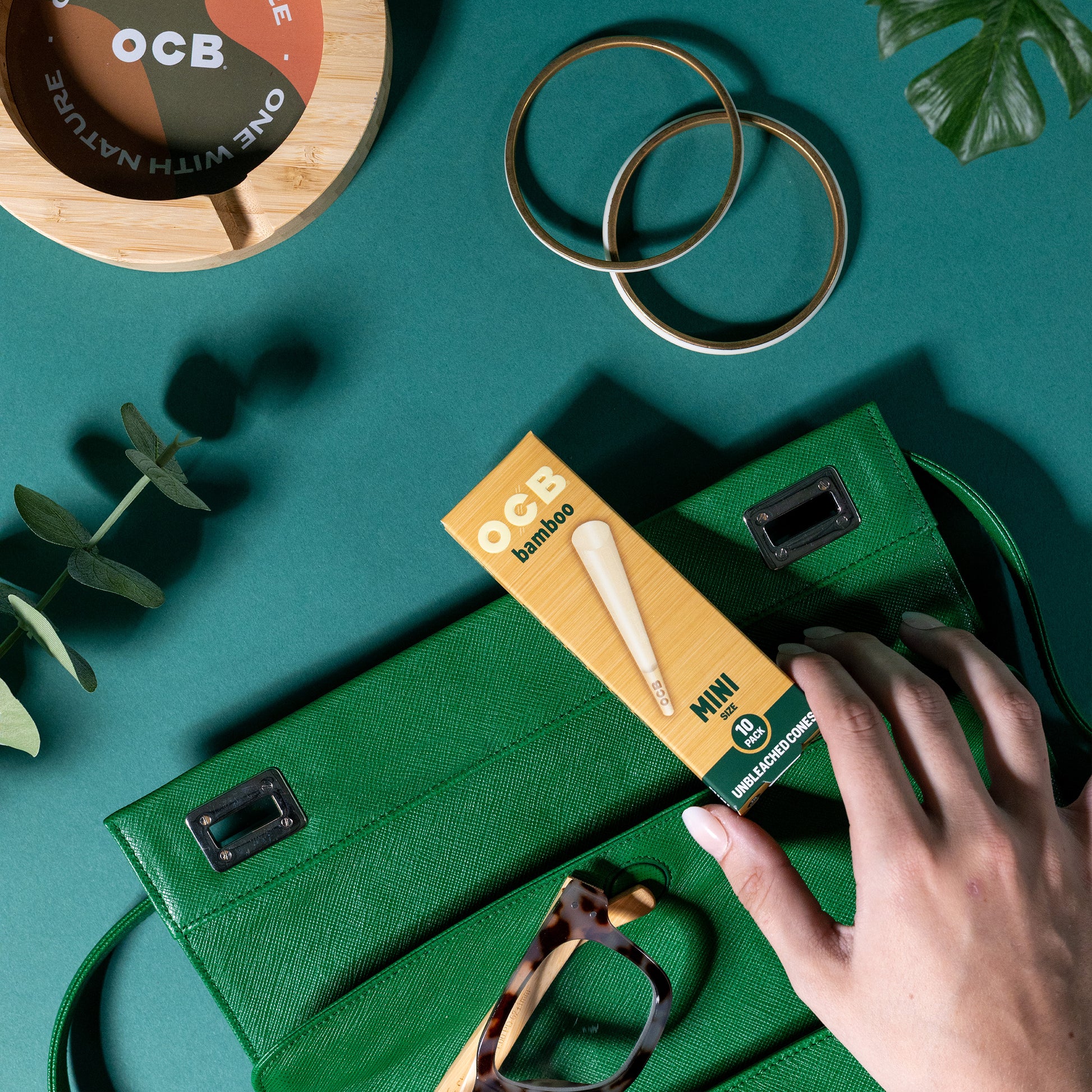 A hand inserts an OCB Bamboo Mini cone pack into a green textured clutch bag, styled on a teal background with gold rings, glasses, and a wooden coaster.