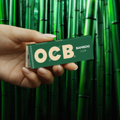 A hand holding a green OCB Bamboo 11/4 rolling paper pack in front of a background of green bamboo stalks.