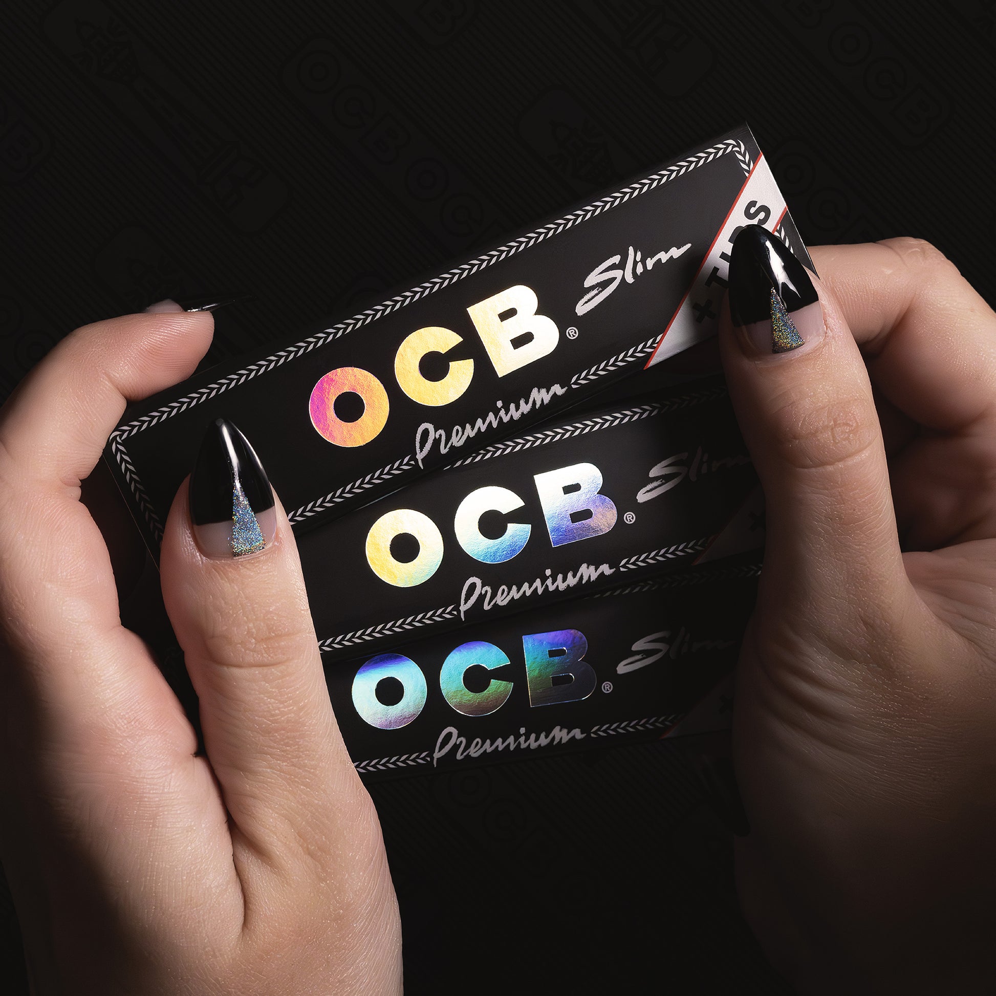 A close-up of two hands holding a slim black OCB Premium Slim package, showing the holographic rainbow logo and white script text against a dark background.