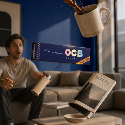 A man in a living room reacts with a surprised expression as floating objects include a coffee mug splashing coffee, a Slim OCB Ultimate booklet, a lighter, and a magazine on a table. The background shows a blue wall and a gray sofa.