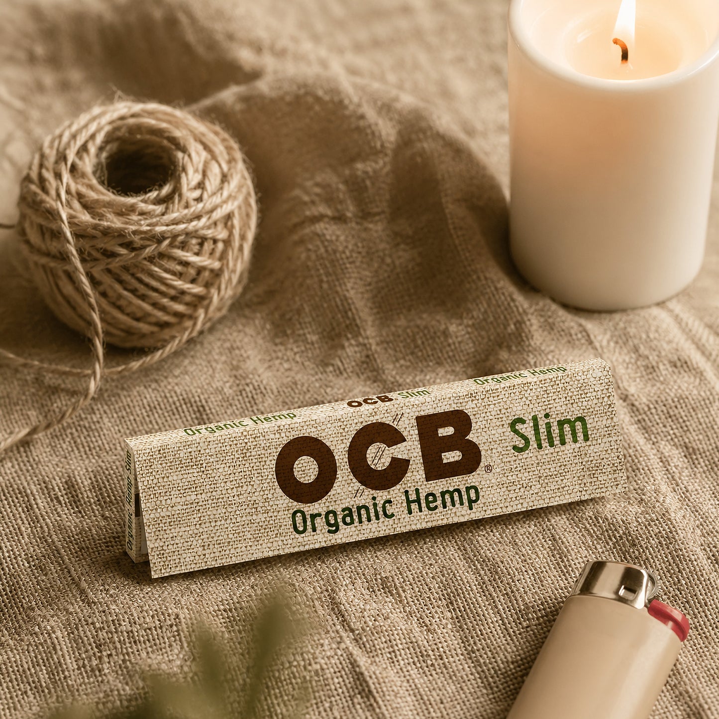 A pack of OCB Slim Organic Hemp rolling paper lies on a rustic burlap surface, styled with a spool of twine, a lit white candle, and a beige lighter.