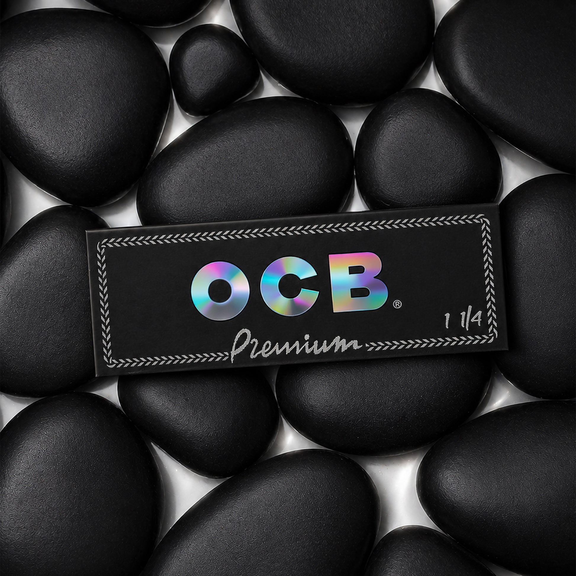 A rectangular black OCB Premium 1 1/4 booklet placed on top of smooth black stones. The OCB logo appears in iridescent rainbow lettering, with 'Premium' and '1 1/4' printed below in white.