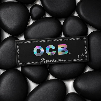 A rectangular black OCB Premium 1 1/4 booklet placed on top of smooth black stones. The OCB logo appears in iridescent rainbow lettering, with 'Premium' and '1 1/4' printed below in white.