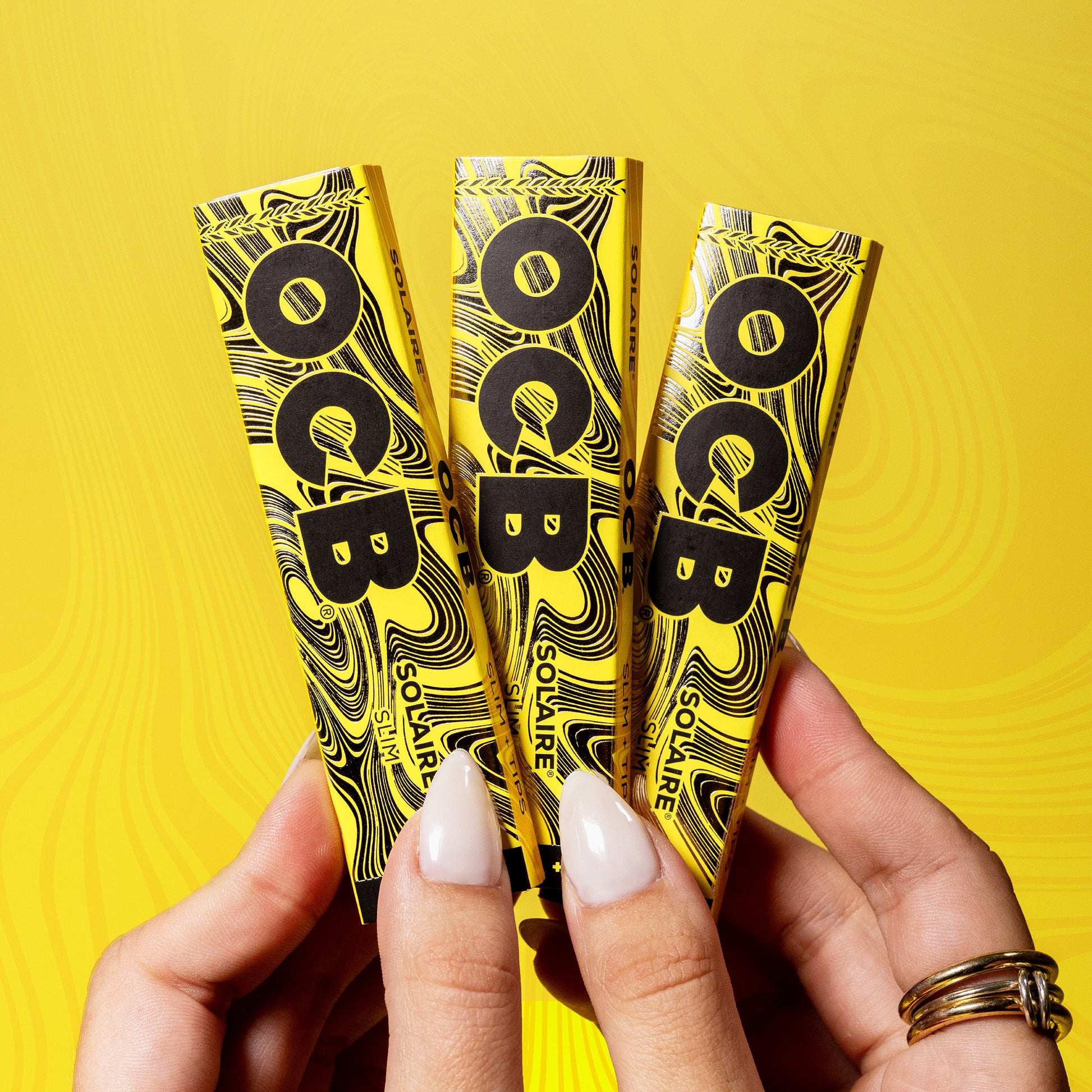 Three yellow and black Soliare rolling paper booklets held in two hands against a bright yellow background with a subtle swirling pattern.