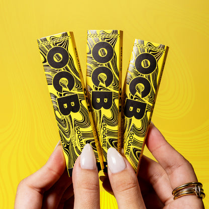 Three yellow and black Soliare rolling paper booklets held in two hands against a bright yellow background with a subtle swirling pattern.